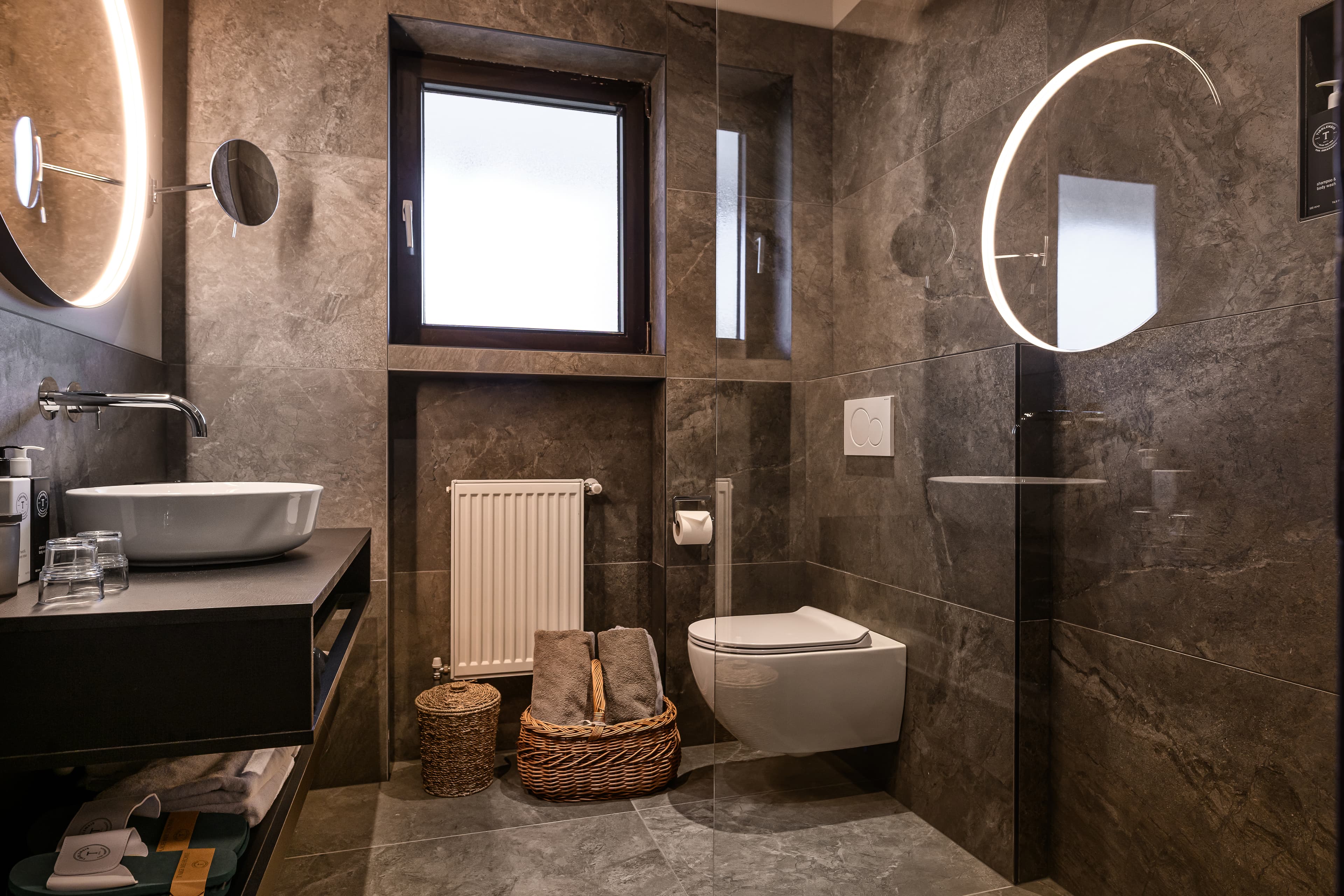 Bathroom with grey stone tiles and round backlit mirror - Hotel Tirolensis